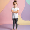 Young girl wearing a white t-shirt and dark leggings standing against a colorful abstract background.