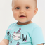 Baby wearing a light blue shirt with a cat unicorn design on a plain background