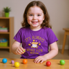 Child wearing a purple shirt with a message, sitting at a table with colorful balls.