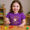 Child wearing a purple shirt with a message, sitting at a table with colorful balls.