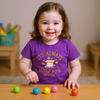 Child wearing a purple shirt with a message, sitting at a table with colorful balls.