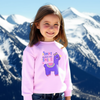 Young girl wearing a pink sweater with a llama graphic and text, standing in front of snowy mountains.
