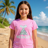 Young girl wearing a pink 'Mermaid' t-shirt on a beach with palm trees and ocean in the background.